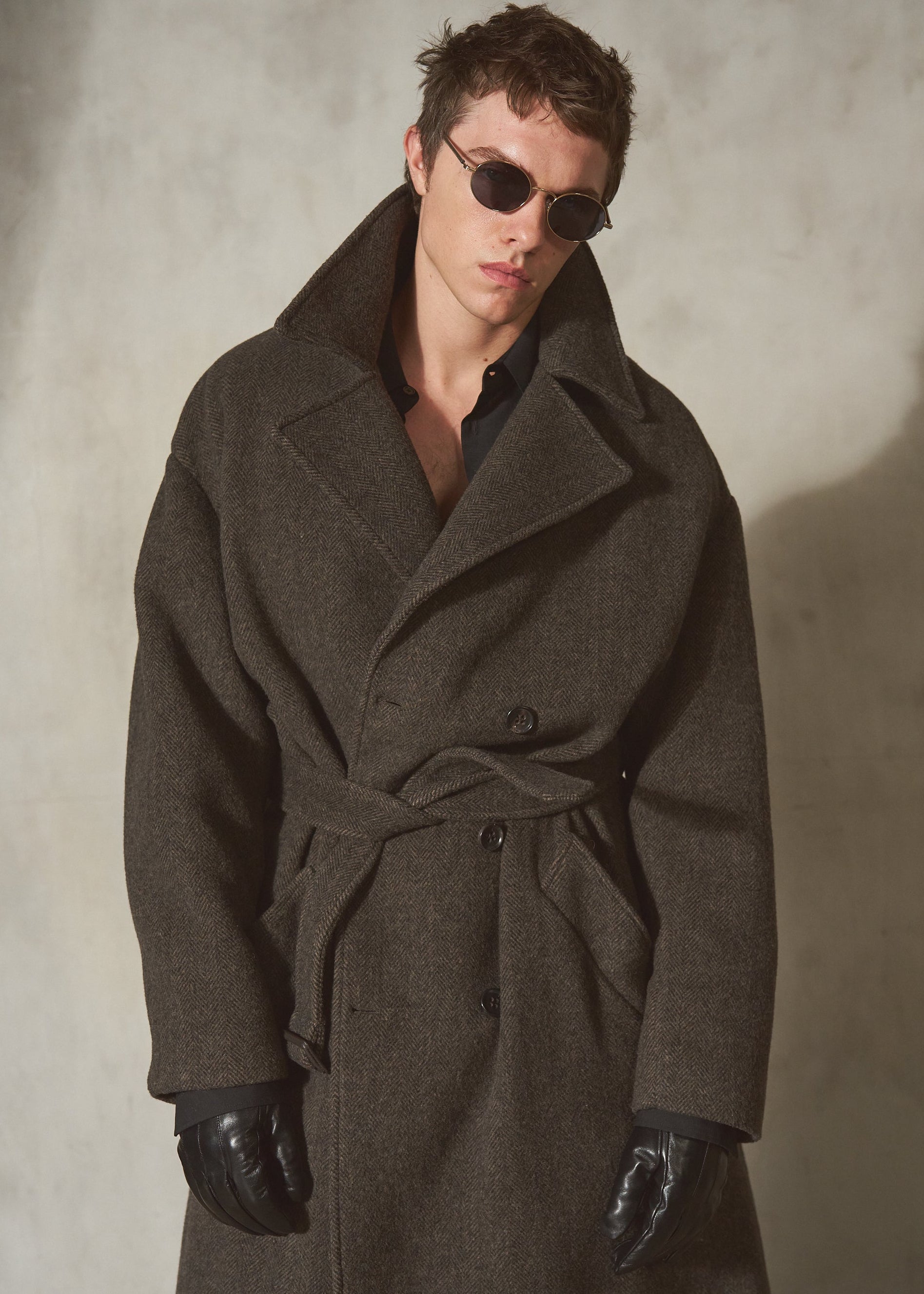 A man wearing a dark brown herringbone wool-blend belted overcoat, black leather gloves, and round sunglasses, standing against a textured light gray wall.