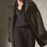 A model wearing a long, oversized dark brown wool-blend coat over a black button-down shirt and black trousers, accessorized with black gloves and dark sunglasses, set against a textured concrete wall.