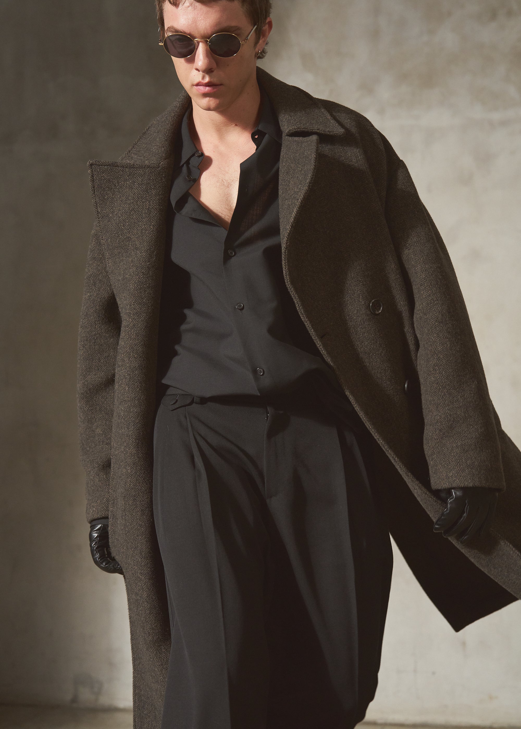 A model wearing a long, oversized dark brown wool-blend coat over a black button-down shirt and black trousers, accessorized with black gloves and dark sunglasses, set against a textured concrete wall.