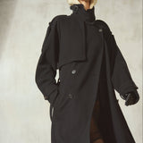 A model wearing a long black trench coat, black baseball cap, and black gloves, standing against a textured light gray wall.
