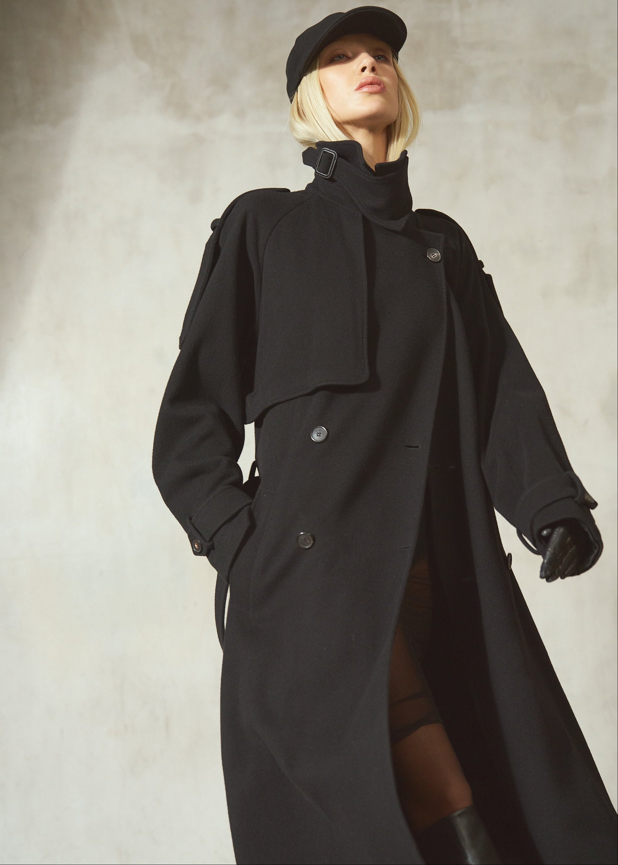 A model wearing a long black trench coat, black baseball cap, and black gloves, standing against a textured light gray wall.
