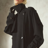 A model wearing a black double-breasted trench coat, black gloves, and a black cap, posing against a textured light gray background.