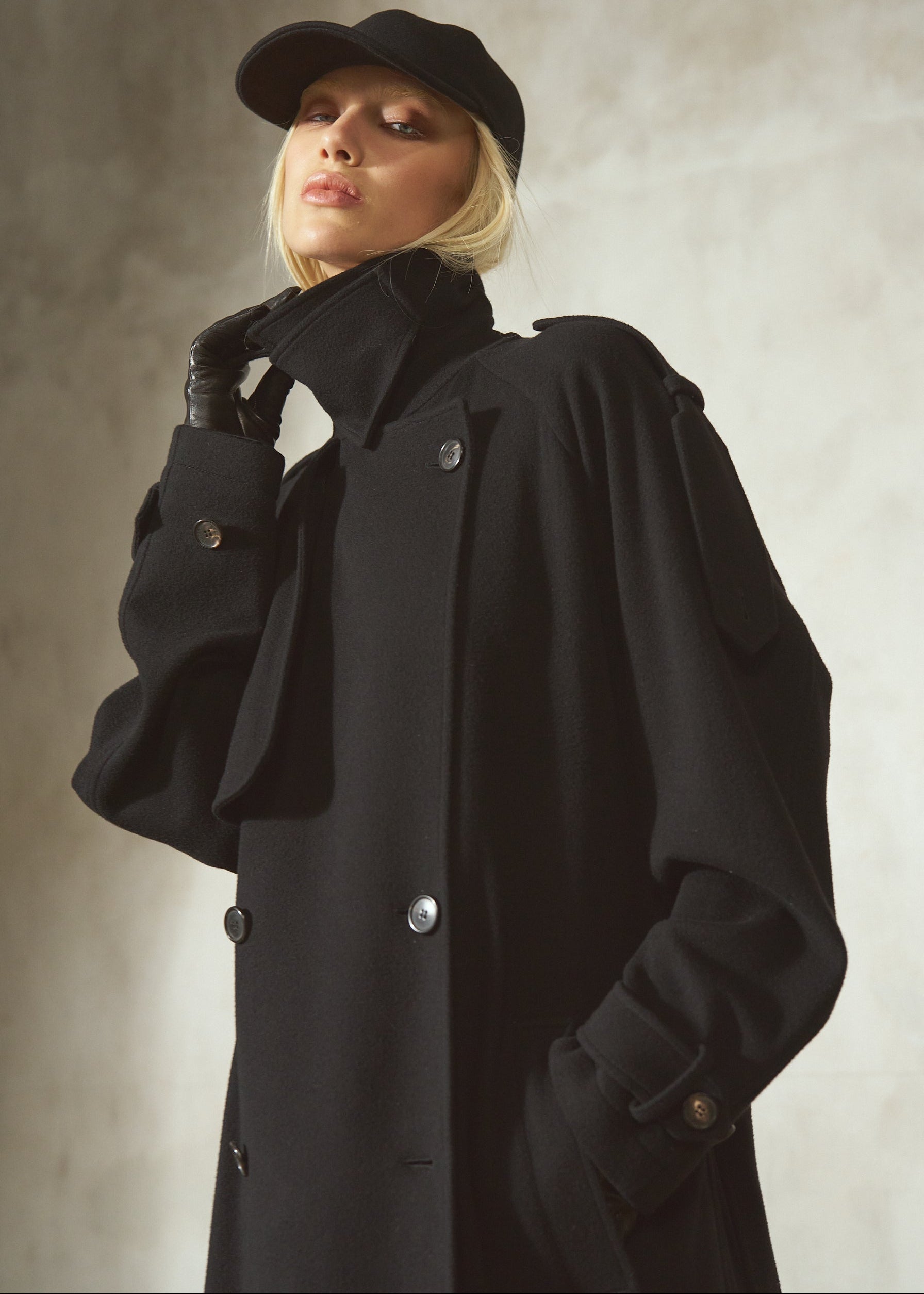 A model wearing a black double-breasted trench coat, black gloves, and a black cap, posing against a textured light gray background.