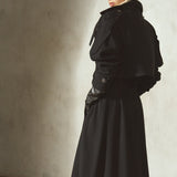 A model wearing a black trench coat, long black skirt, black leather gloves, black pointed-toe boots, and a black baseball cap, standing in profile with a textured concrete wall.