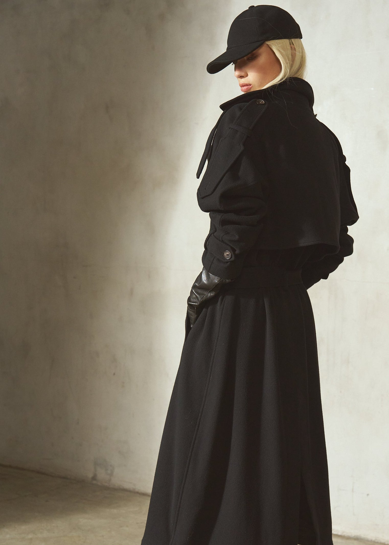 A model wearing a black trench coat, long black skirt, black leather gloves, black pointed-toe boots, and a black baseball cap, standing in profile with a textured concrete wall.