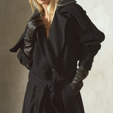 A model wearing a black cashmere trench coat with structured shoulders, a matching black belt, black leather gloves, and a black baseball cap, posed against a textured light gray wall.