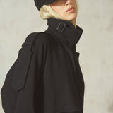A model wearing a black high-collar coat with a shoulder strap detail, a black cap, and blonde hair, posed against a textured light gray wall.