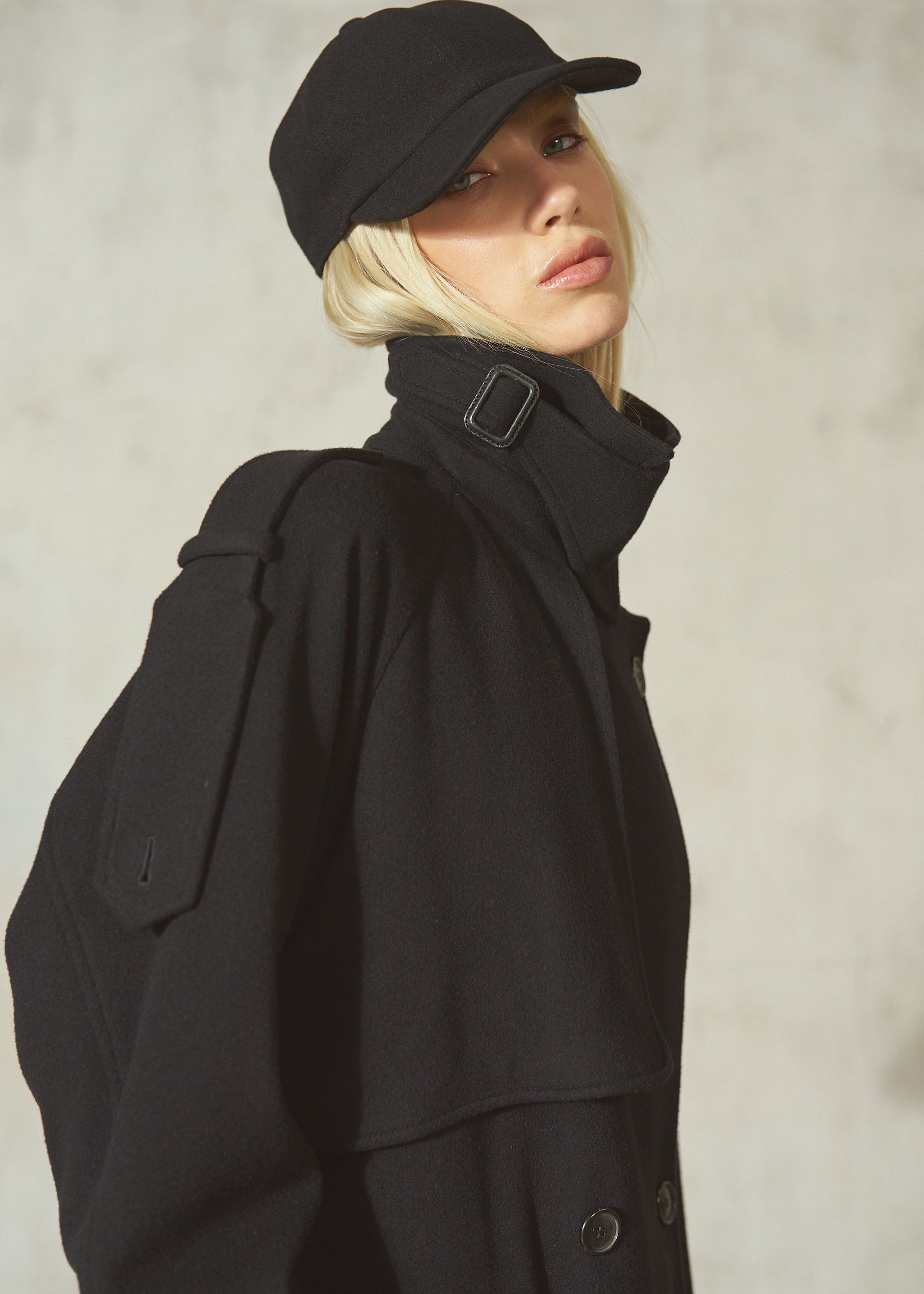 A model wearing a black high-collar coat with a shoulder strap detail, a black cap, and blonde hair, posed against a textured light gray wall.