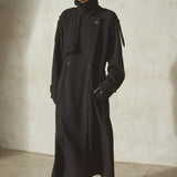 A model wearing a long black trench coat, black baseball cap, black gloves, and black boots, standing against a textured concrete wall.