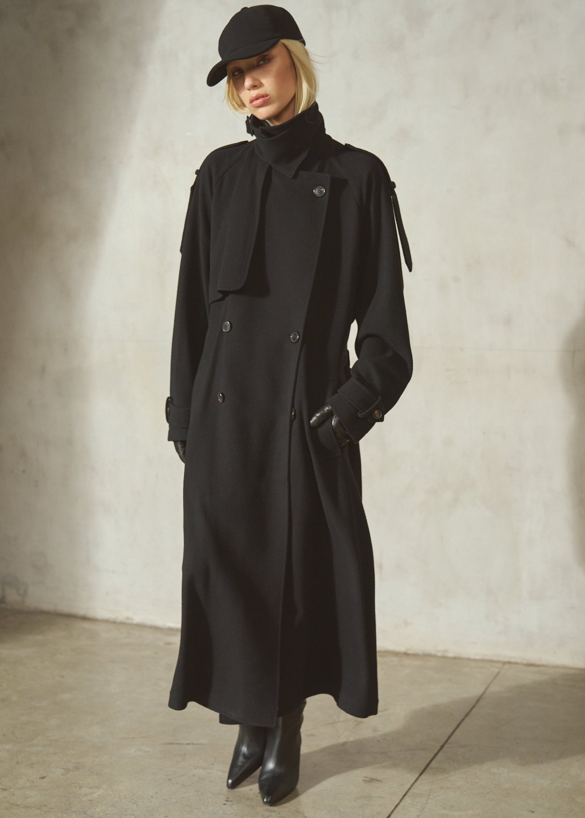 A model wearing a long black trench coat, black baseball cap, black gloves, and black boots, standing against a textured concrete wall.