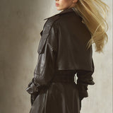 Dressed in a dark brown leather trench coat with round sunglasses, a blonde woman poses in a minimalist gray environment.