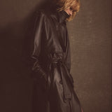 A blonde woman styled in a dark brown leather trench coat, photographed in a minimalist gray environment.