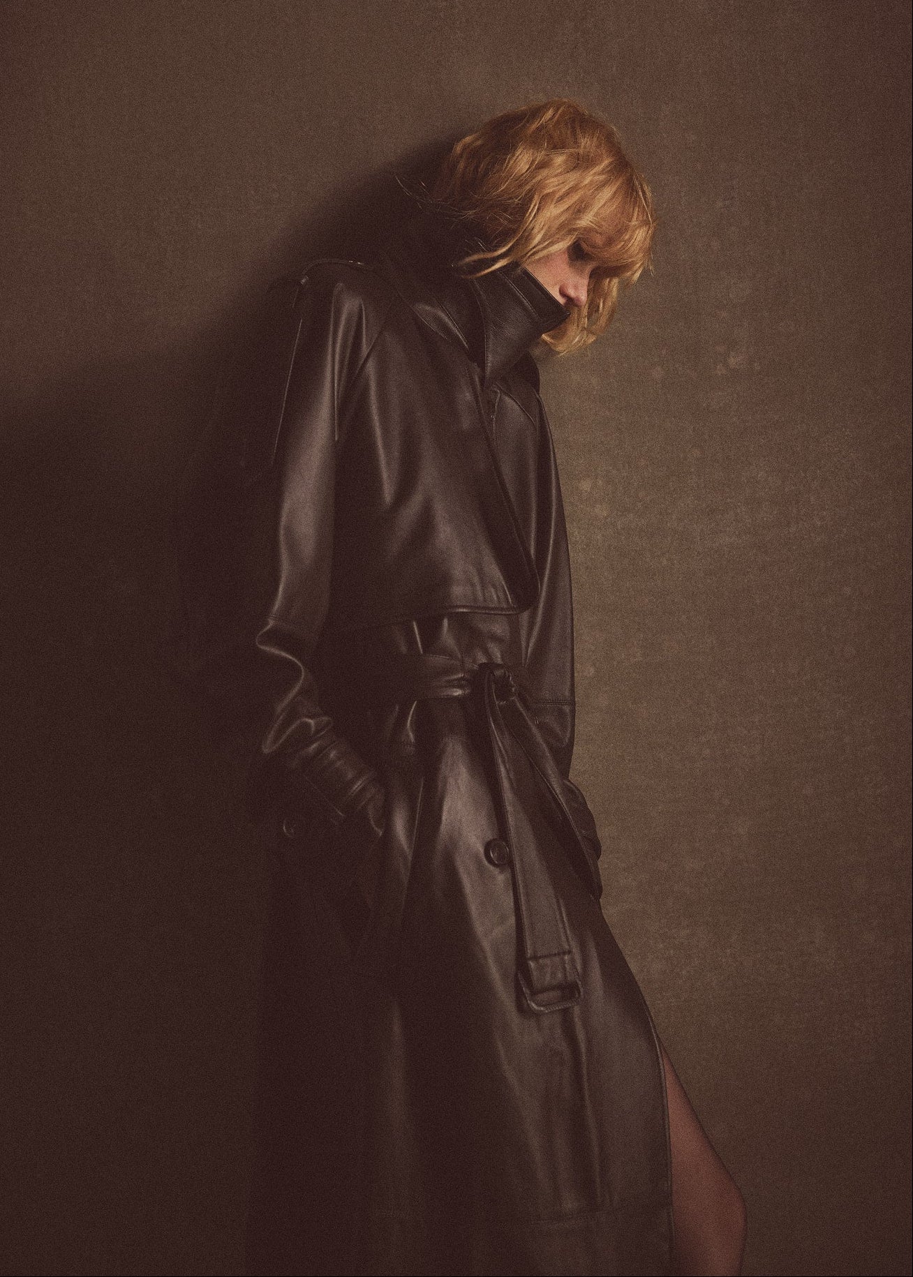 A blonde woman styled in a dark brown leather trench coat, photographed in a minimalist gray environment.