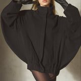 A model wearing a dramatic oversized black coat with wide cape-like sleeves, black gloves, and a black cap, posed against a textured gray wall.