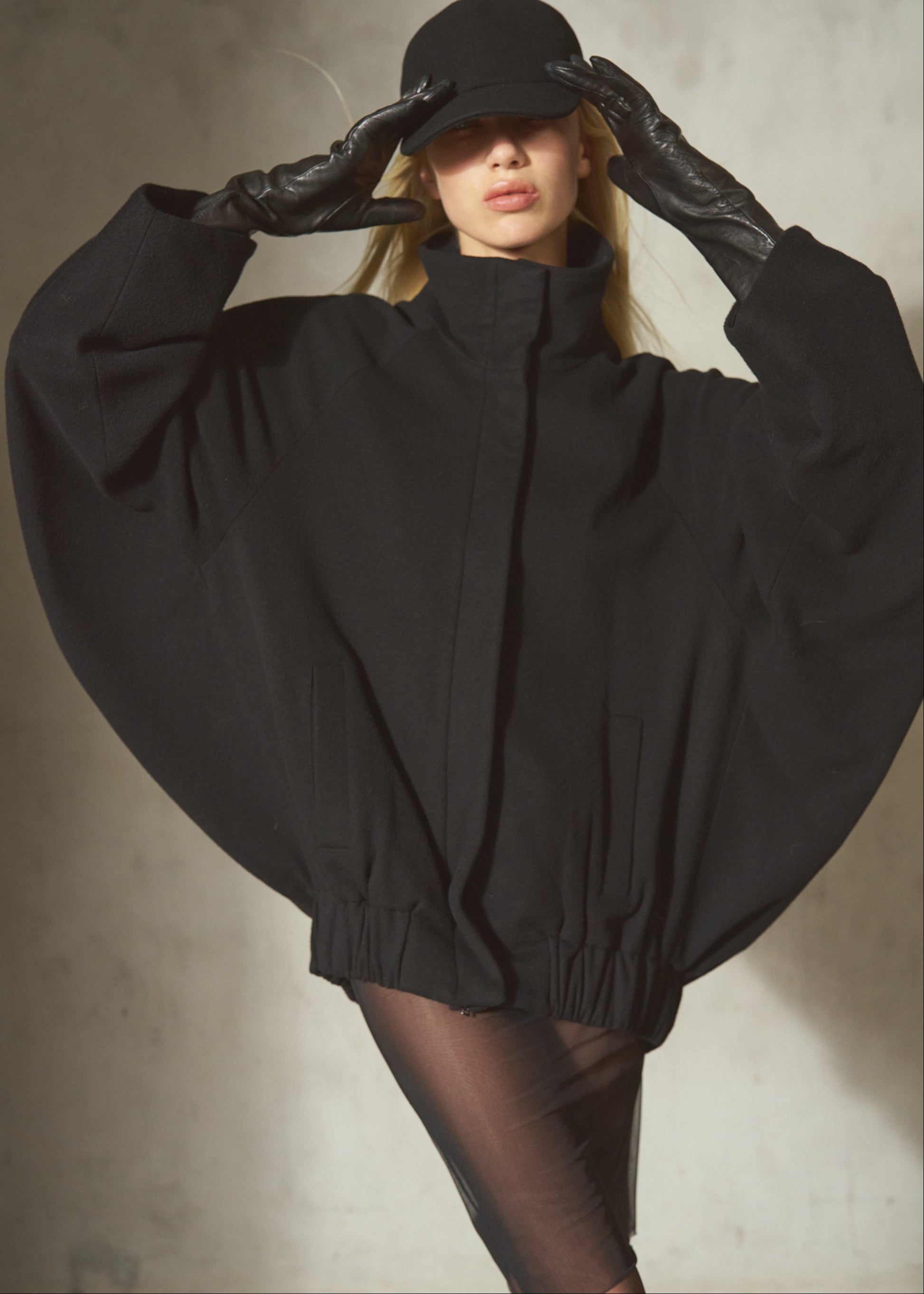 A model wearing a dramatic oversized black coat with wide cape-like sleeves, black gloves, and a black cap, posed against a textured gray wall.