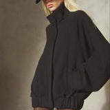 A model wearing an oversized black zip-up jacket with a high collar and elastic waist, paired with a black baseball cap, black tights, and posed against a textured gray concrete wall.