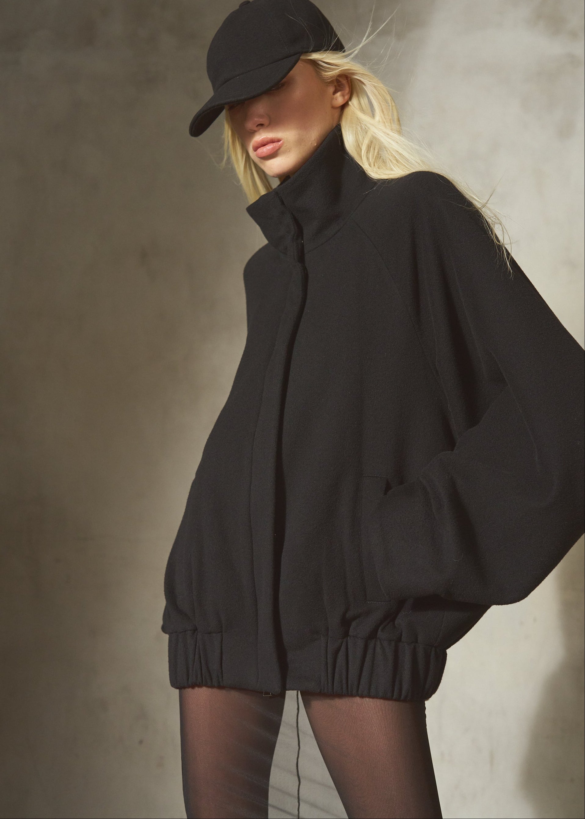 A model wearing an oversized black zip-up jacket with a high collar and elastic waist, paired with a black baseball cap, black tights, and posed against a textured gray concrete wall.