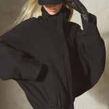 A model wearing a black oversized zip-up jacket with batwing sleeves, a black baseball cap, and a black glove, posing on a textured concrete wall.