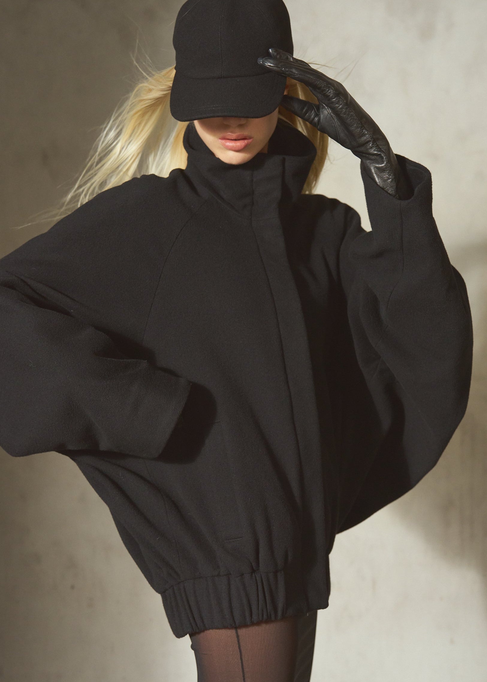 A model wearing a black oversized zip-up jacket with batwing sleeves, a black baseball cap, and a black glove, posing on a textured concrete wall.