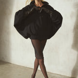 A model wearing a black oversized batwing-sleeve jacket, sheer black tights, black strappy heels, and a black baseball cap, standing infront a textured gray wall.