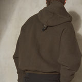 A male model viewed from behind wearing an oversized dark olive hooded jacket and dark trousers, standing against a textured concrete wall.