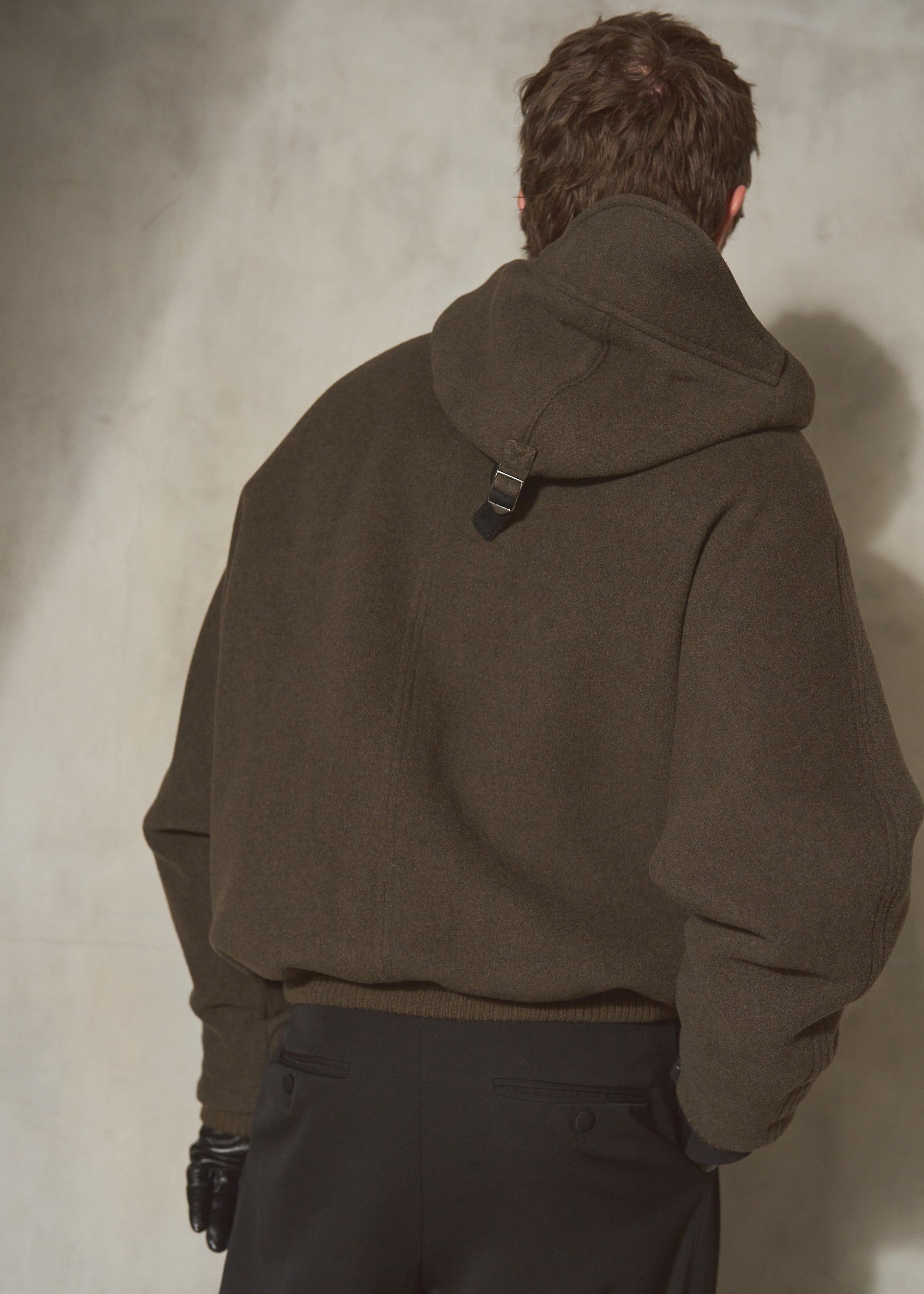 A male model viewed from behind wearing an oversized dark olive hooded jacket and dark trousers, standing against a textured concrete wall.