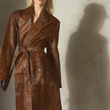 A woman wearing a brown and black leopard-print coat with a matching tie belt, a black baseball cap, and black gloves, standing against a textured gray wall.