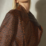 A close-up lifestyle shot of a model wearing a brown and black leopard pony hair coat, and a black wool cap against a textured light gray wall.