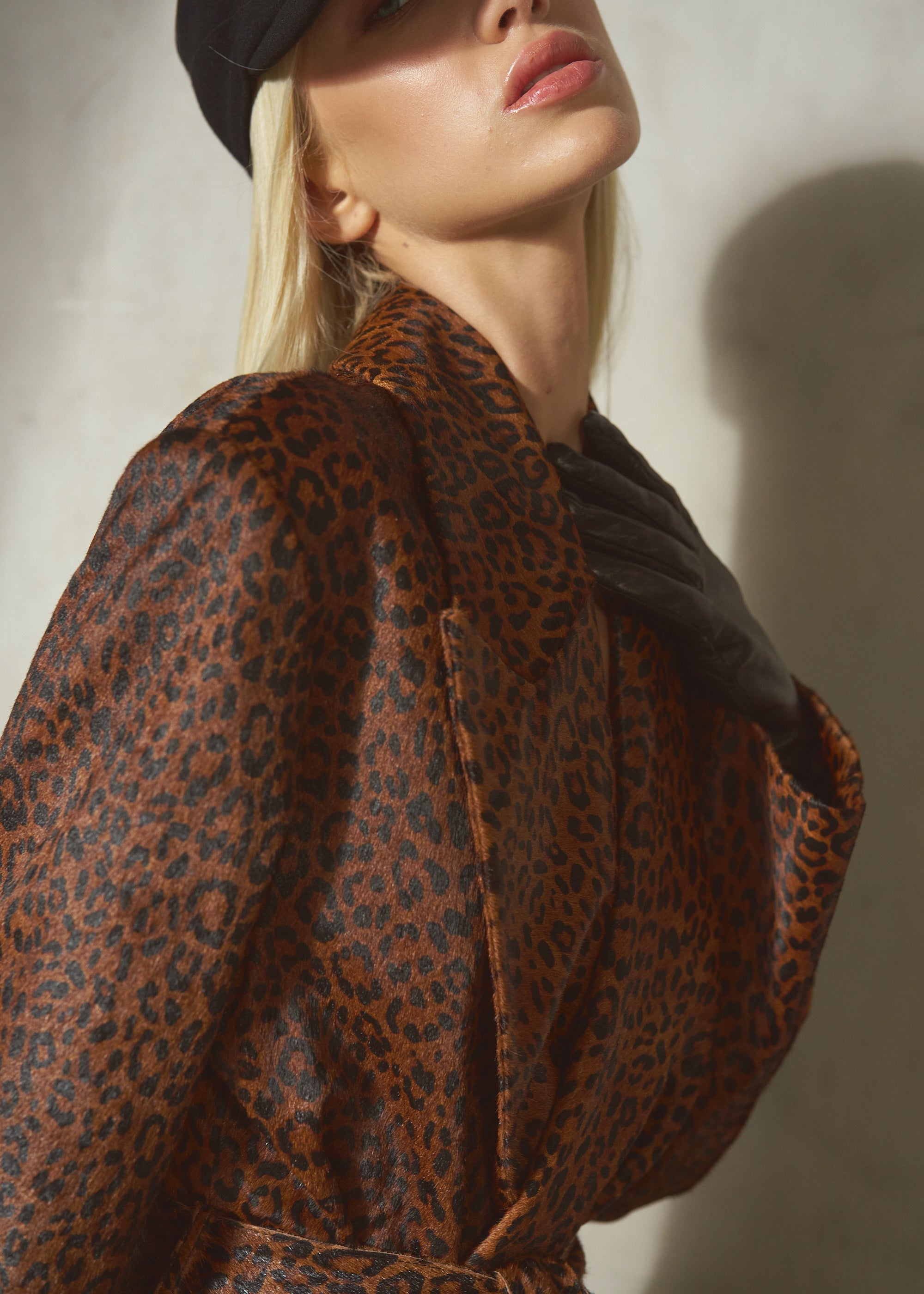 A close-up lifestyle shot of a model wearing a brown and black leopard pony hair coat, and a black wool cap against a textured light gray wall.