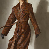 A model wearing a brown and black leopard-print topcoat with a matching waist belt, black gloves, and a black baseball cap, standing against a textured concrete wall with a strong shadow.