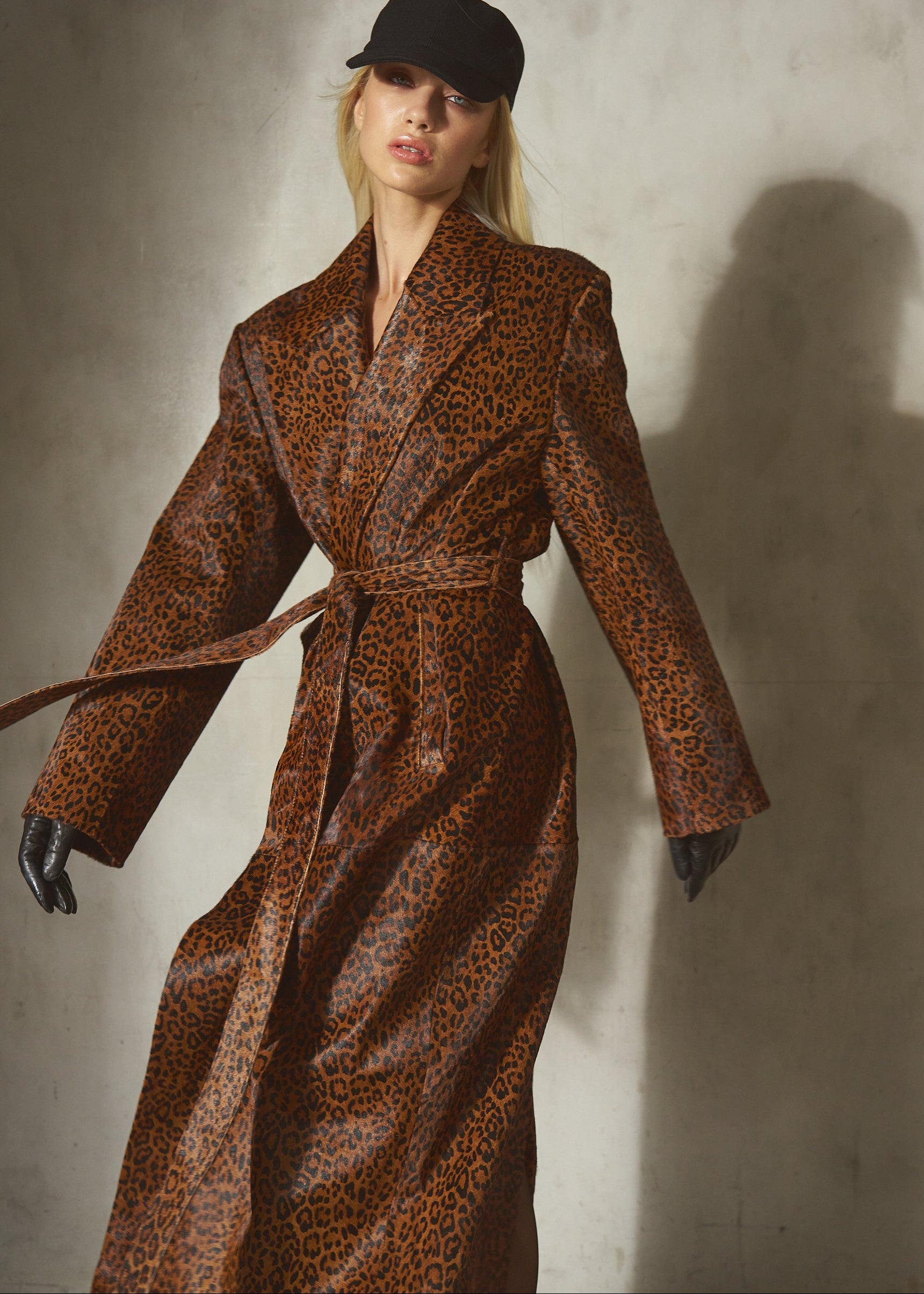 A model wearing a brown and black leopard-print topcoat with a matching waist belt, black gloves, and a black baseball cap, standing against a textured concrete wall with a strong shadow.
