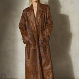 A model stands against a textured concrete wall wearing a long brown and black leopard-print coat, a black cap, and black pointed-toe heels.