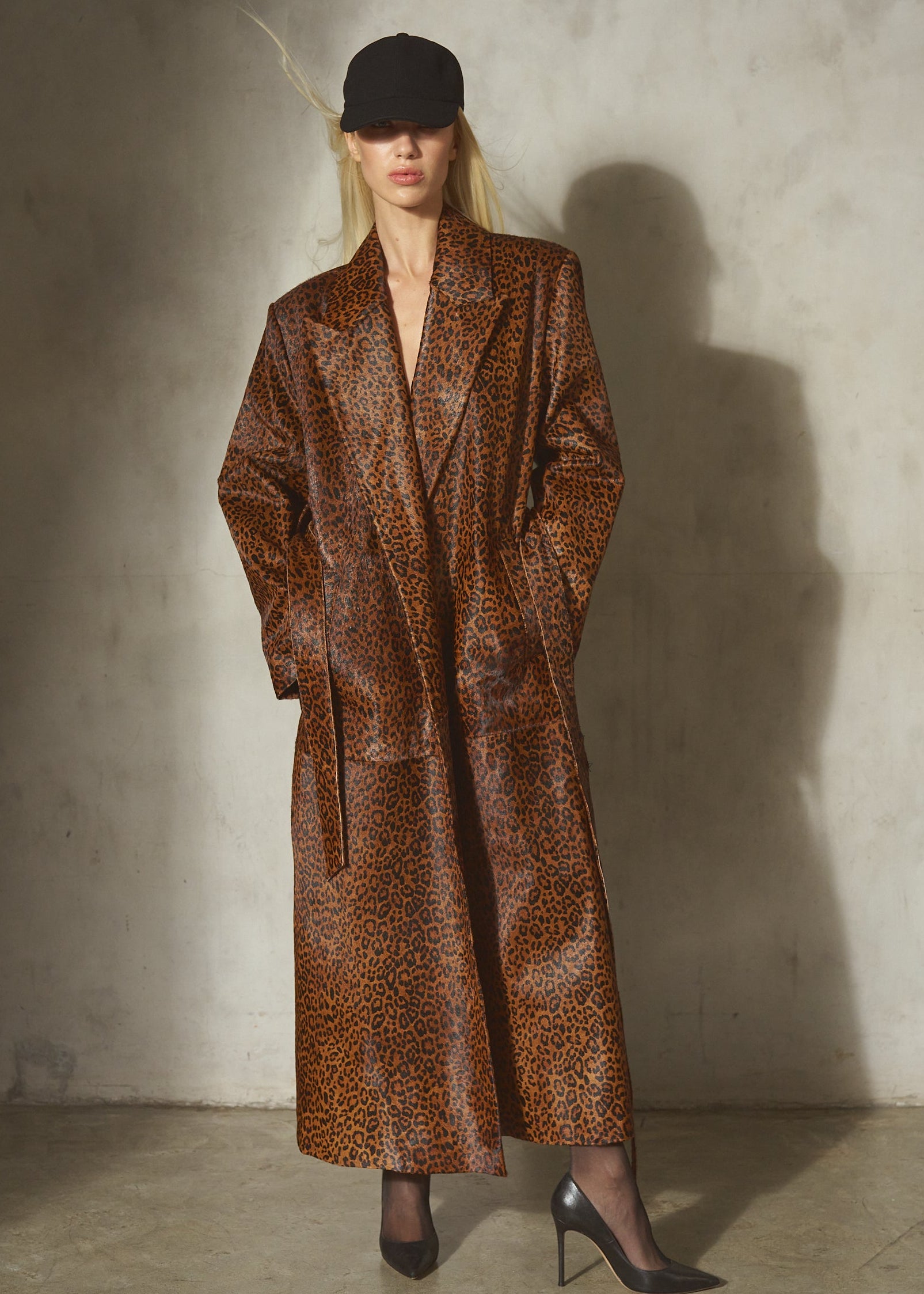 A model stands against a textured concrete wall wearing a long brown and black leopard-print coat, a black cap, and black pointed-toe heels.