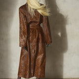 A model with long blonde hair is shown from behind wearing a brown and black leopard-print pony hair long coat, a black baseball cap, and black heels, standing against a textured concrete wall.