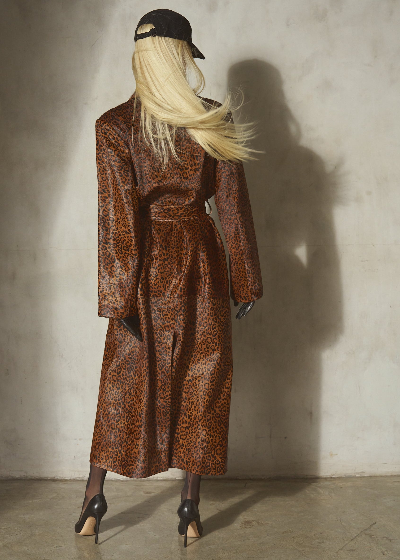 A model with long blonde hair is shown from behind wearing a brown and black leopard-print pony hair long coat, a black baseball cap, and black heels, standing against a textured concrete wall.