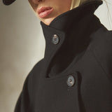 A close-up of a model wearing a black wool coat with a high collar and a matching black baseball cap, against a neutral gray background.