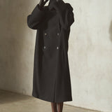 A model wearing a long black double-breasted wool coat, black wool cap, black gloves, and black strappy heels, standing against a textured concrete wall.