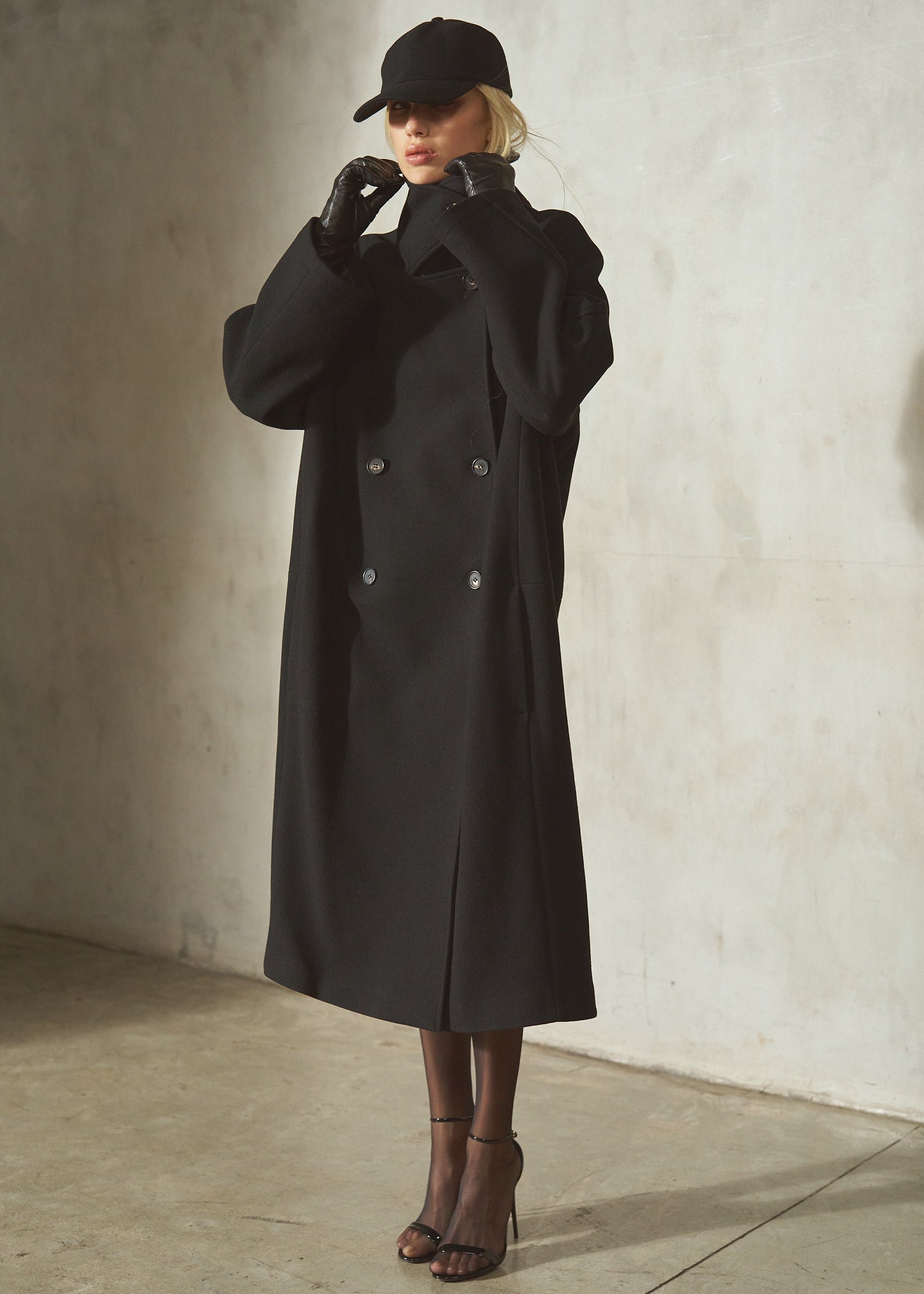 A model wearing a long black double-breasted wool coat, black wool cap, black gloves, and black strappy heels, standing against a textured concrete wall.