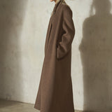 A woman stands in profile wearing a long brown wool-blend coat, black cap, and black pointed heels against a textured concrete wall.