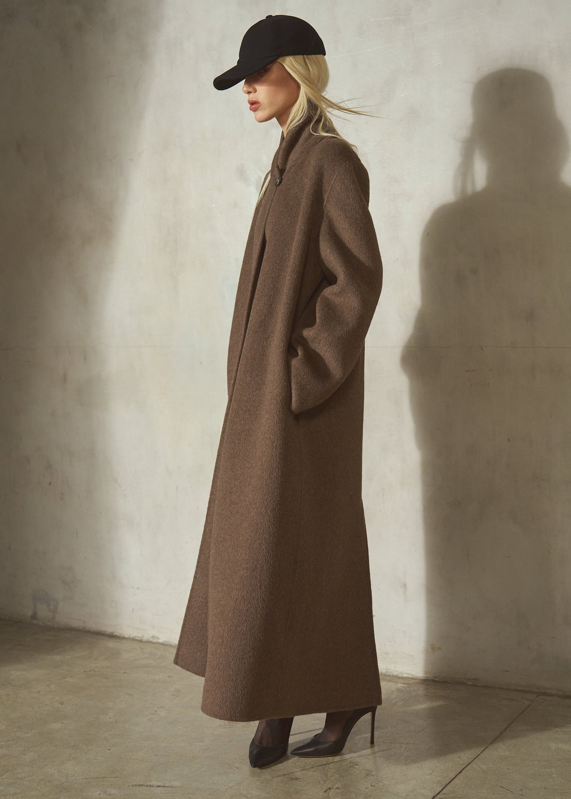 A woman stands in profile wearing a long brown wool-blend coat, black cap, and black pointed heels against a textured concrete wall.
