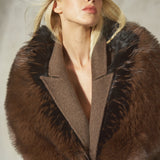 A blonde model wearing a brown mohair coat with a faux fur collar and a black baseball cap, photographed against a neutral gray background.
