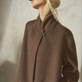 A blonde woman wearing a black baseball cap and a brown wool long coat with a high collar and single button closure, posed against a textured light gray wall.