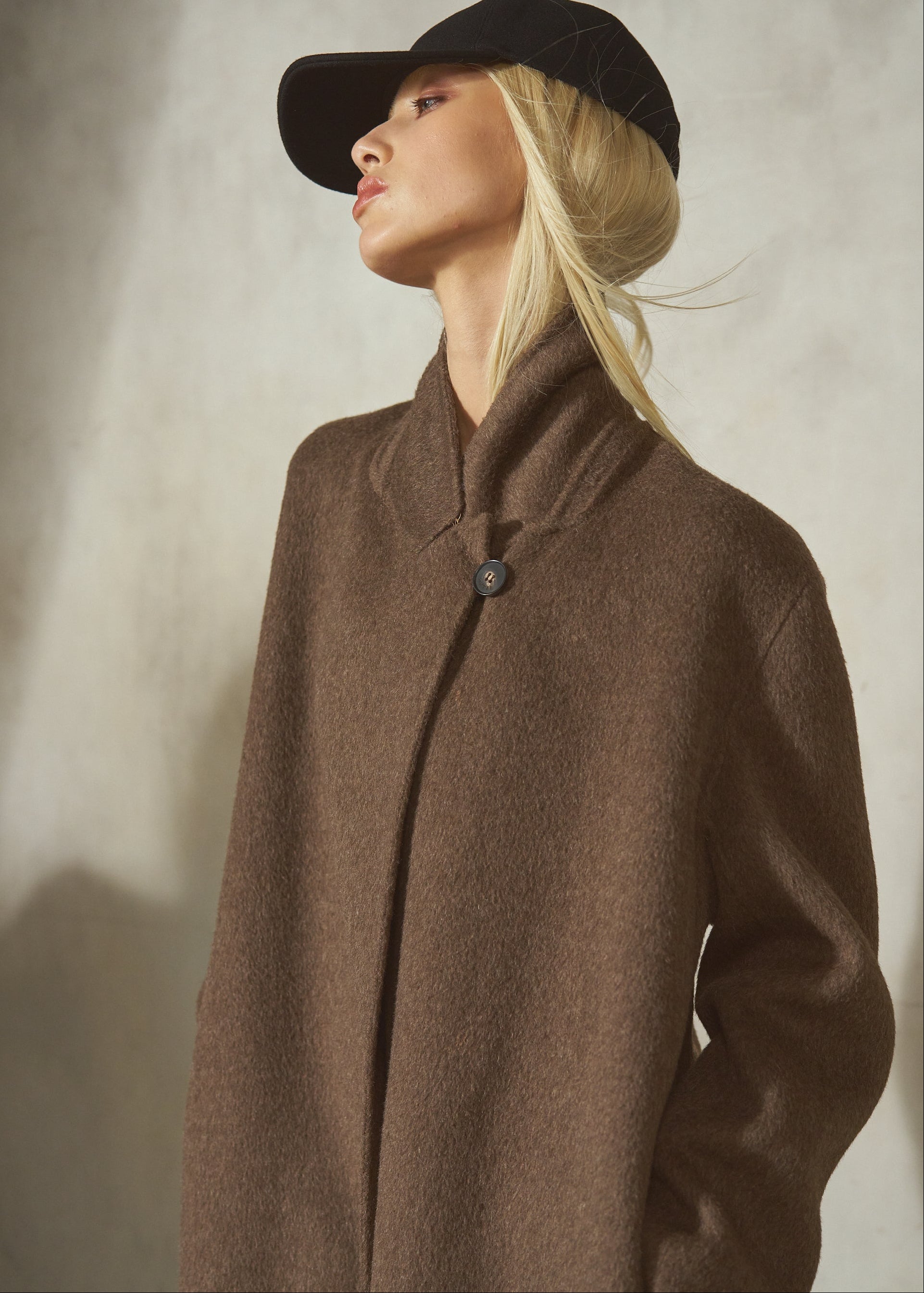 A blonde woman wearing a black baseball cap and a brown wool long coat with a high collar and single button closure, posed against a textured light gray wall.