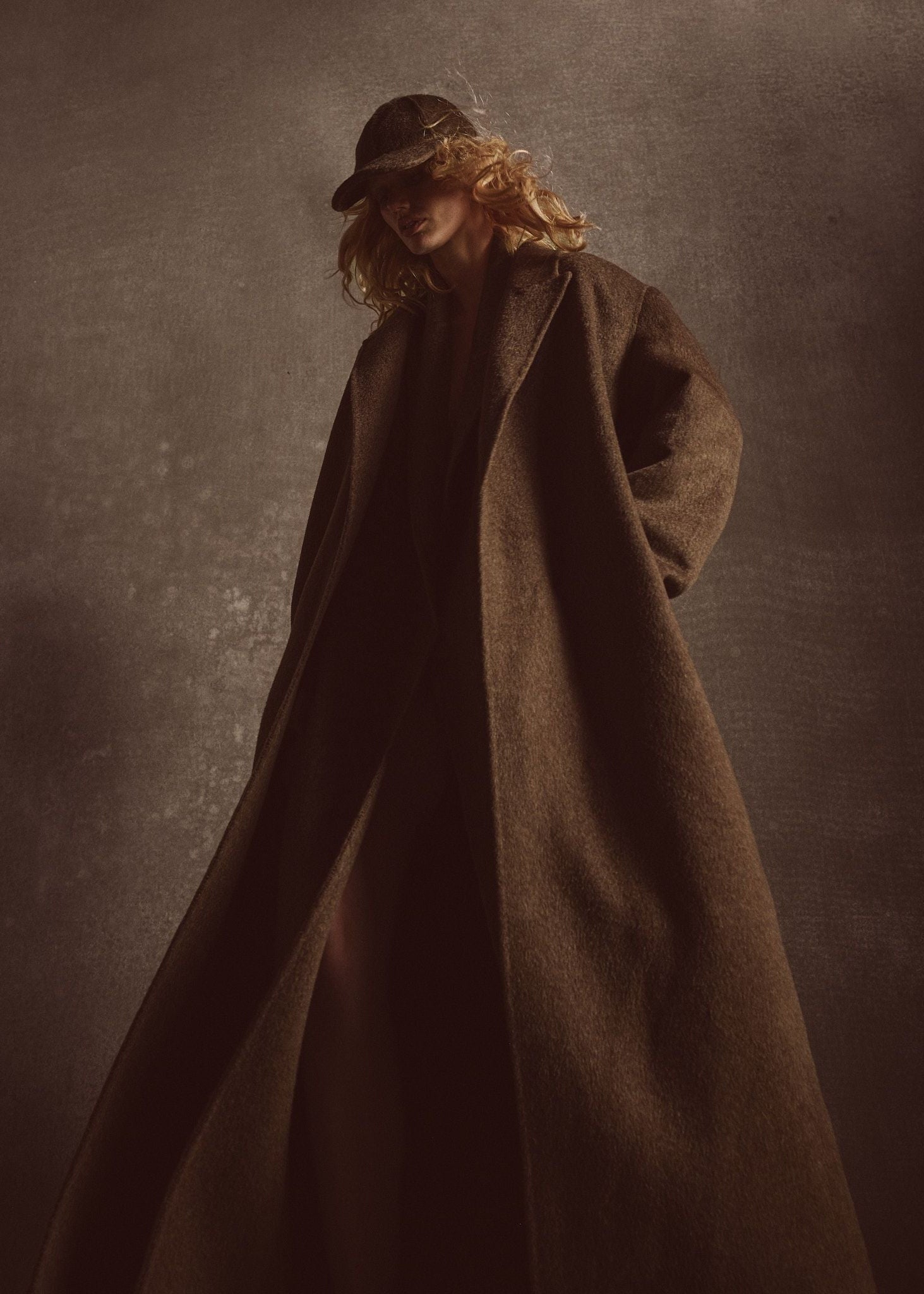 Model wearing a long brown coat and hat against a dark background