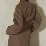 A model wearing a brown wool coat with a matching belt, a black baseball cap, standing against a textured light gray wall.