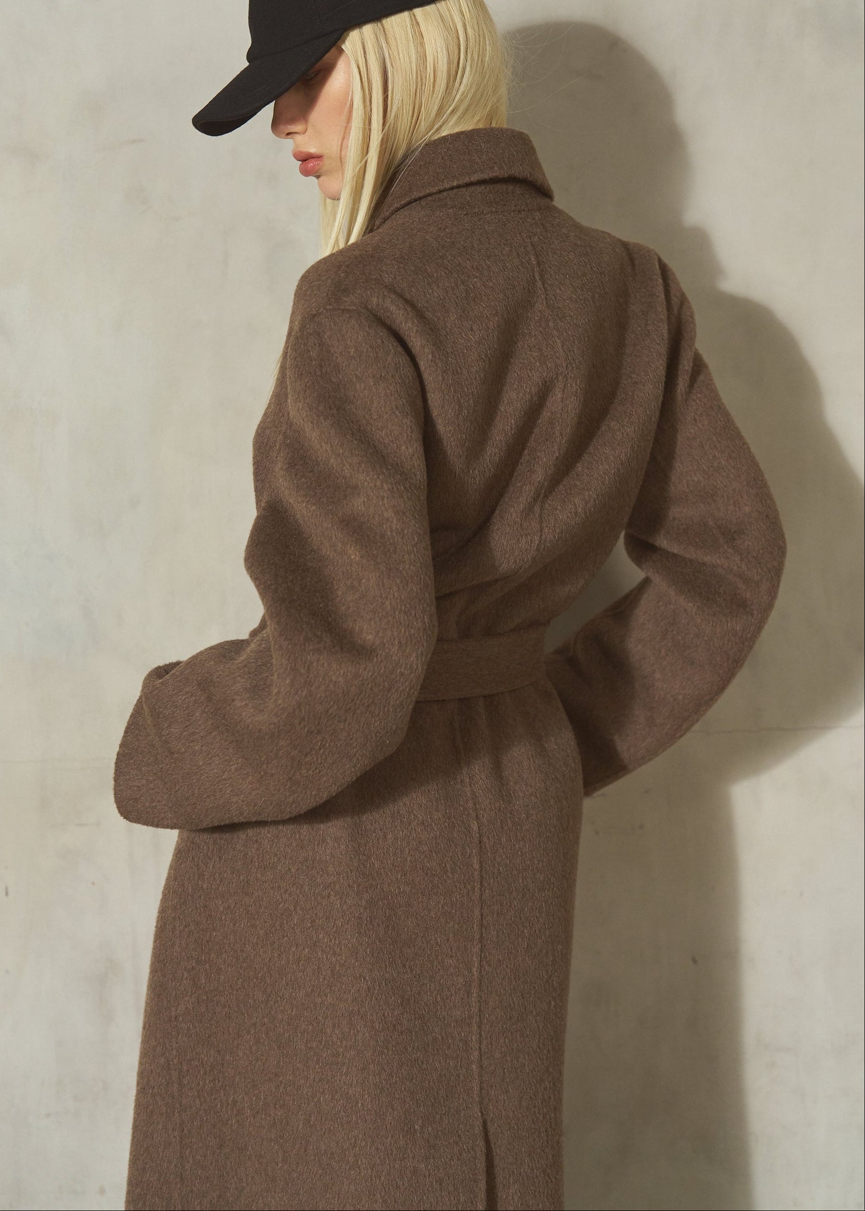 A model wearing a brown wool coat with a matching belt, a black baseball cap, standing against a textured light gray wall.
