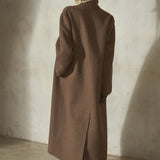 A blonde hair model stands with her back to the camera wearing a long brown topcoat, black cap, and black high heels against a textured concrete wall.