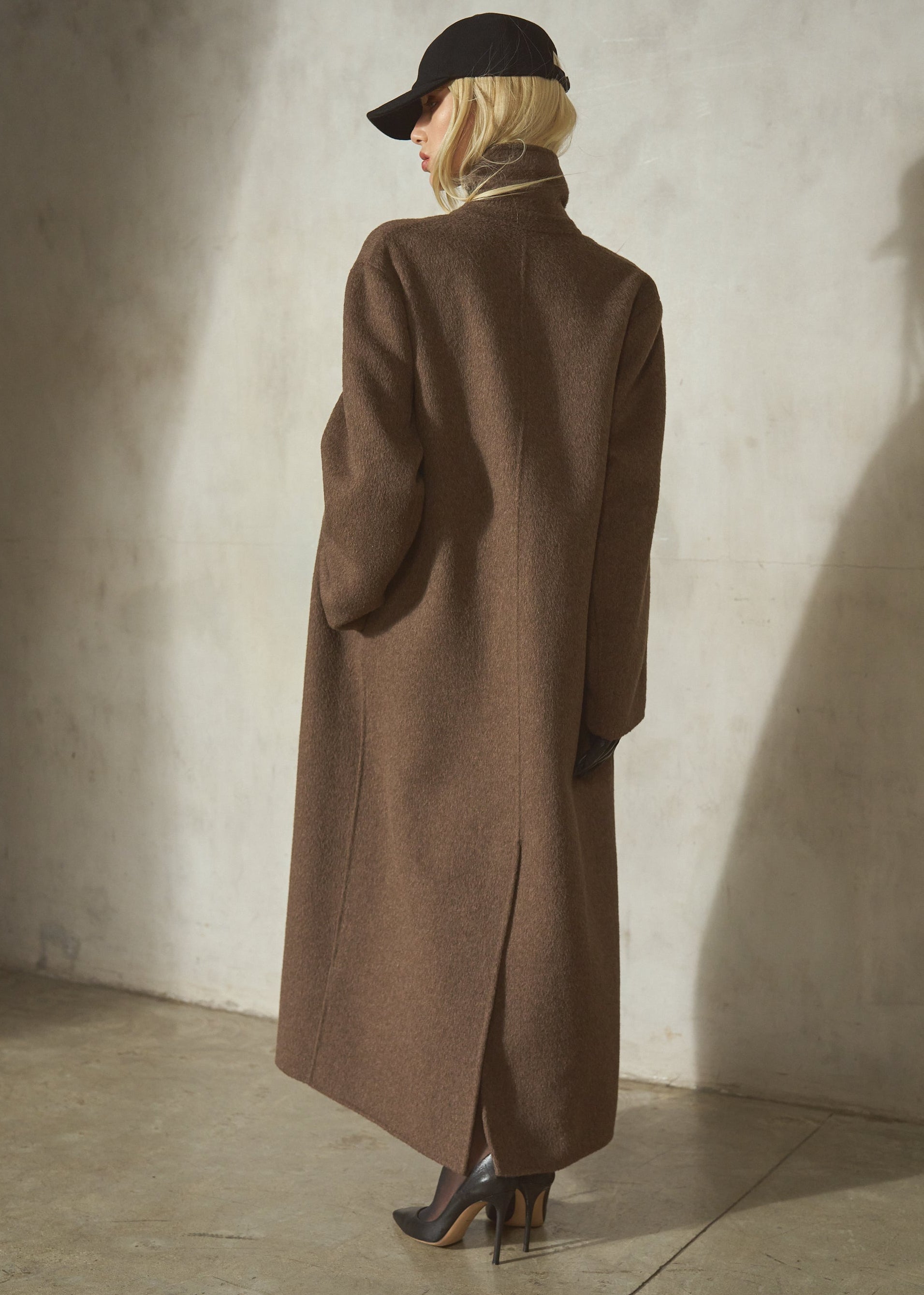 A blonde hair model stands with her back to the camera wearing a long brown topcoat, black cap, and black high heels against a textured concrete wall.