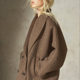A blonde model wearing a brown double-breasted textured coat and sunglasses, photographed from the side against a textured gray studio background.