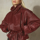 A model wearing a belted oversized dark red ponyhair fur jacket with a high neck, paired with black pants and a black baseball cap.
